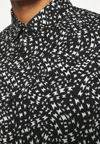 Camicia nera con un motivo a zigzag bianco che si attorciglia, colletto con bottoni e una texture di tessuto liscia. Sono visibili diversi bottoni.