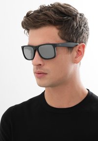 Sunglasses med en matt svart ram, fyrkantig form och gråa linser. Har en logotyp på sidan och en slät yta.