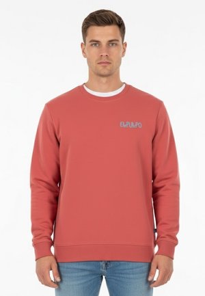 Hombre vestido con un sudadera color salmón que lleva el texto "ELPULPO" en el pecho, combinada con jeans de color azul claro, de pie frente a un fondo blanco.