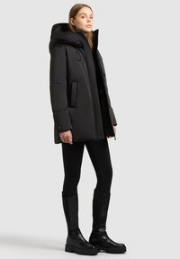 Femeie îmbrăcată cu o haină cu glugă neagră și umplutură, leggings negre și cizme negre până la genunchi, stând într-un fundal simplu, deschis la culoare.
