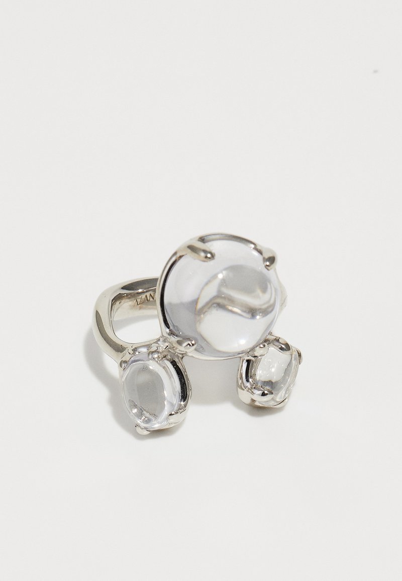 Sudraba gredzens ar trim skaidrām ovālām formām akmeņiem, gludām virsmām, novietots uz eleganta, pulēta apļa.