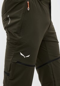 Παντελόνια υπαίθρια σκούρου πράσινου χρώματος με τσέπες με φερμουάρ, πορτοκαλί έλξη φερμουάρ, ετικέτα Durastretch και λευκό στυλιζαρισμένο λογότυπο στο μηρό.