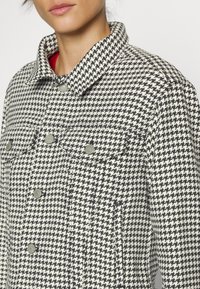 Džínová bunda s černobílým houndstooth vzorem, límcem, patentkami a dvěma náprsními kapsami. Texturovaná látka, přiléhavý střih.