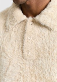 Maglietta beige pelosa con colletto, caratterizzata da quattro bottoni e un tessuto morbido e testurizzato. Il colore dei bottoni corrisponde a quello della maglietta.