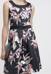 Květinové šaty v černé tkanině s růžovými, modrými a zelenými akcenty. Mají výstřih do lodičky, přiléhavý pas a rozšířenou sukni.