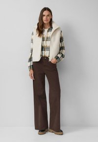 Off-White Fleece-Weste über einem karierten Flanellhemd in Grün, Gelb und Weiß; braune weit geschnittene Hose; schwarze Plateau-Stiefel.