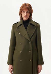 Dubbelknäppt olivegrön ullrock med bred krage, strukturerat tyg, dekorativa knappar och sidofickor. Rektangulär form.