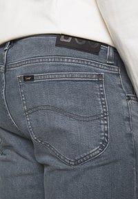 Grå denimjeans med klassisk femficksdesign, med en svart läderlapp i midjan och subtila sömdetaljer på bakfickorna.