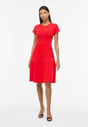 Femme debout portant une robe rouge vif à manches courtes, longueur genou, avec une jupe à volants, et des talons noirs ouverts, sur un fond blanc.