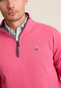 Roze fleece pullover met een half-rits ontwerp, voorzien van een logo patch van een zeilboot en contrasterende donkerblauwe accenten op de kraag.