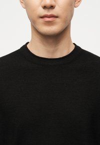 Gros plan sur une personne portant un pull ras du cou noir uni, montrant les lèvres, le menton, le cou et le haut de la poitrine sur un fond neutre.