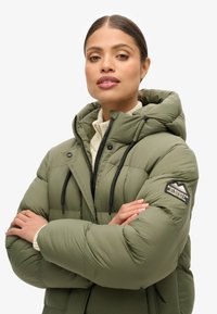 Veste doudoune verte avec capuche, présentant un motif matelassé, des cordons de serrage noirs et une patte de logo sur la manche. Texture de tissu douce.