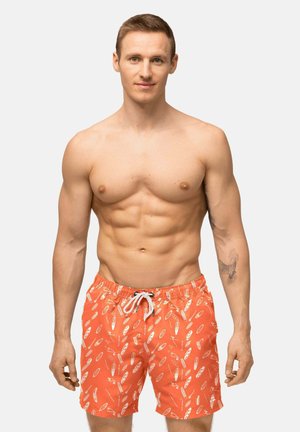 Uomo muscoloso con capelli castani corti che indossa pantaloncini da bagno arancioni con motivi bianchi a forma di pagaia, in piedi davanti a uno sfondo chiaro e uniforme.