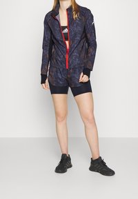Marineblå og brun mønstret sportjakke med rød lynlås, parret med sorte shorts og sorte sneakers; stoffet er glat med et tætsiddende design.