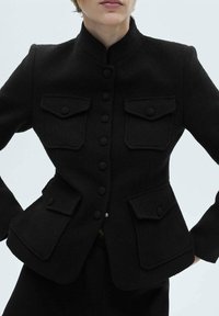 Veste noire texturée avec col mandarin, dotée de six boutons sur le devant et de deux poches poitrine avec rabats. Coupe ajustée avec une silhouette structurée.