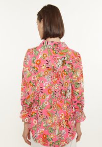 Blusa a maniche lunghe in tessuto rosa con un vivace motivo floreale accentuato da dettagli verdi e arancioni. Include un colletto e maniche arricciate.