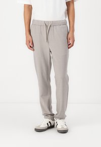 Persoon in lichtgrijze sweatpants met een trekkoord in de taille en witte sneakers met zwarte strepen, staand tegen een witte achtergrond.