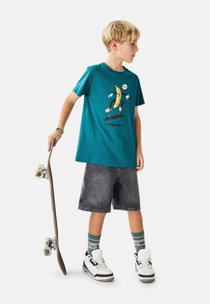 Tyrkis bomuld T-shirt med en tegneserie-banan grafik, kombineret med sorte denimshorts og hvide højtop sneaks. Skateboard holdt i venstre hånd.