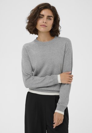 Rövid barna hajú nő szürke hosszú ujjú pulóvert fehér mandzsettával és fekete nadrággal, egyszerű háttér előtt állva.