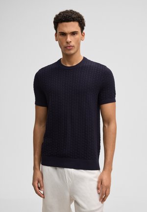 Mann steht und trägt einen dunkelblauen, strukturierten Pullover mit kurzen Ärmeln und hellbeige Hosen vor einem schlichten grauen Hintergrund.