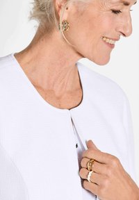 Weiß strukturierte Jacke mit rundem Ausschnitt, ausgestattet mit einem Knopfverschluss. Gold- und weiße Ringe an den Fingern, die geschichtete Accessoires präsentieren.
