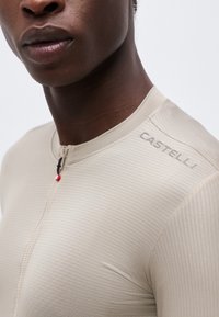 Detailný záber na osobu nosiacu béžový rebrovaný cyklistický dres s malým predným zipsom a logom "CASTELLI" na ramene.