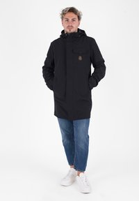 Zwarte regenjas met capuchon, voorzien van een borstzak en ritssluiting. Gecombineerd met blauwe spijkerbroeken en witte sneakers.