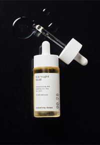 Przezroczysta szklana butelka z białą zakrętką z pipetą, zawierająca żółty nawilżający olejek. Etykieta zawiera informacje o produkcie w czarnym tekście.