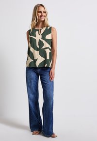 Bezrukávý zeleno-bežový top s abstraktním vzorem, s malým detailem ve tvaru klíčové dírky, v kombinaci s modrými širokými džínami a světlými sandály.