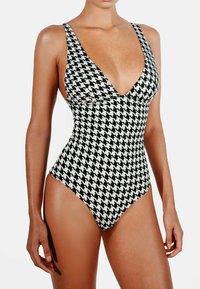 Jednodílné plavky s černobílým vzorem houndstooth, s hlubokým V-výstřihem, širokými ramínky a vysokými výřezy na nohách. Hladká textura látky.