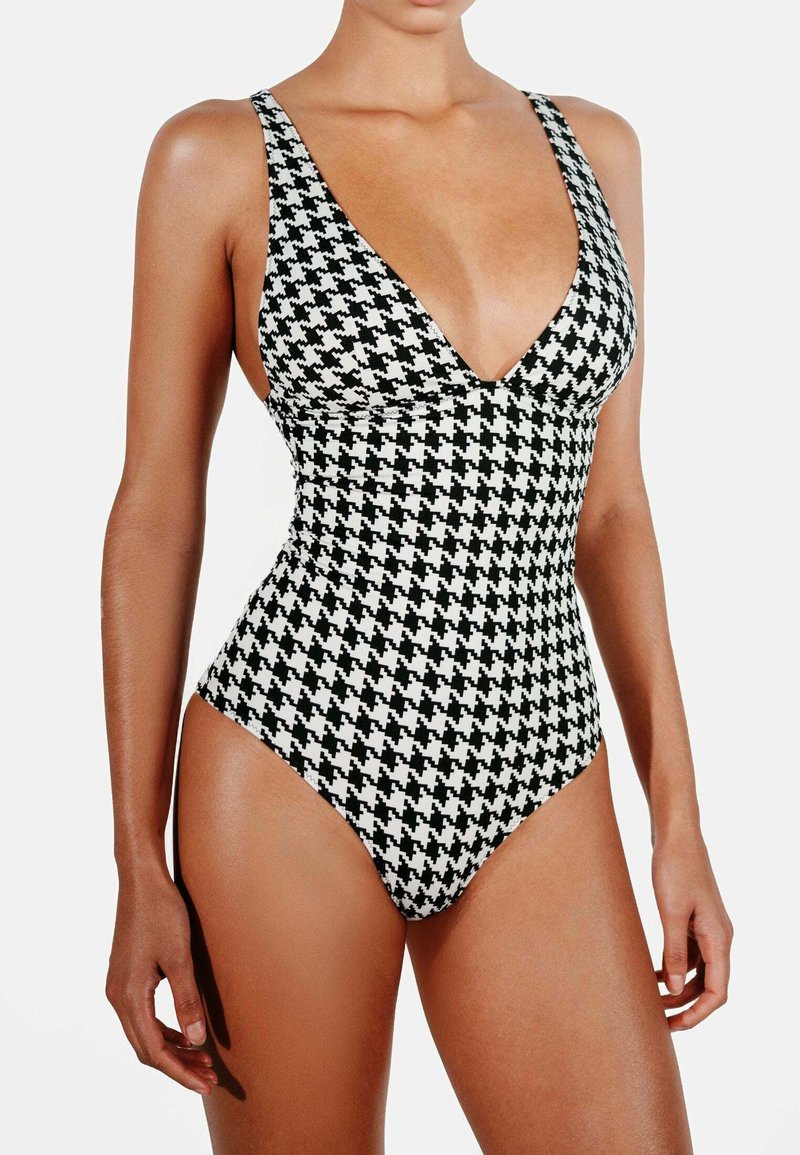 Jednodílné plavky s černobílým vzorem houndstooth, s hlubokým V-výstřihem, širokými ramínky a vysokými výřezy na nohách. Hladká textura látky.