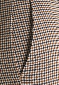 Brązowa, czarna i biała tkanina w wzór houndstooth z teksturowanym splotem, prezentująca dopasowany kształt i szczegóły szwów.