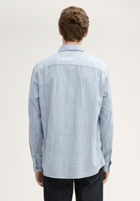 Lichtblauwe langemouwen shirt van textuurstof. Klassieke kraag, met knopen afgewerkte manchetten, en een ronde zoom. Achteraanzicht dat de zijnaden toont.