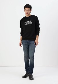 Fekete pulóver "KARL LAGERFELD" logóval, kék farmerral és fekete csizmával viselve. Egyszerű dizájn, bordázott mandzsetták és gallér.