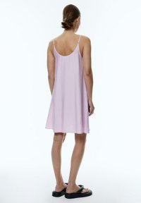 Rosa ärmelloses Kleid mit lockerer Passform, hergestellt aus leichtem Stoff, mit einem runden Rückenausschnitt und einem fließenden Saum. Dazu werden schwarze flache Sandalen getragen.