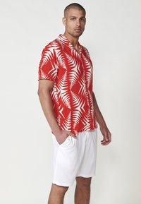 Kurze Ärmel, rotes Hemd mit weißem Farnmuster; kombiniert mit weißen Shorts. Das Hemd hat eine Knopfleiste und einen lässigen Schnitt.