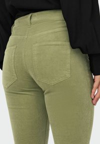 Pantalon en velours côtelé vert avec poches arrière, design ajusté et tissu texturé. La taille est haute, complétant la silhouette générale.