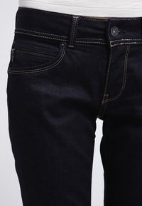 Detailní záběr na tmavě modré džíny s béžovým švem, kapsou na zip a knoflíkovým zapínáním, nošené s bílou košilí.