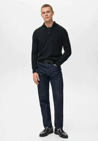 Schwarze Zip-Up-Jacke, dunkelblaue Jeans, schwarzer Gürtel und schwarze Schuhe. Mann steht vor einem weißen Hintergrund, die Hände in den Taschen, mit neutralem Gesichtsausdruck.
