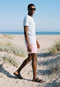 Homme portant des lunettes de soleil, un t-shirt blanc, un short rose et des sandales noires marchant sur une plage de sable avec des herbes de dunes et l’océan en arrière-plan.