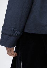 Χέρι χαλαρό στο πλάι, φοράει σκούρο μπλε σακάκι με κουμπωτό λουράκι μανσετών και μαύρη υφασμάτινη φούστα με κάθετες λευκές λεπτομέρειες.