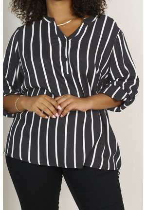 Blouse rayée noir et blanc avec un col en V, détail avec boutons, manches retroussées, en tissu léger, associée à un pantalon noir.