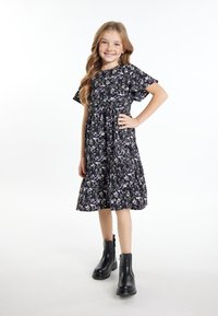 Zwarte bloemenjurk met korte, zwierige mouwen, gerimpelde taille en een rok tot de knie. Draagt zwarte enkellaarzen.