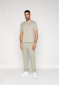 Ανοιχτό μπεζ μπλουζάκι με κεντημένο λογότυπο «Champion», συνδυασμένο με ανοιχτό γκρι παντελόνι cargo με ελαστικές μάνσες και τσέπες, και λευκά αθλητικά παπούτσια.