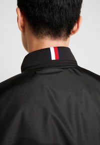 Sort jakke med tekstureret finish, der har en krave fremhævet af røde, hvide og blå striber. Viser bagsiden af nakken.