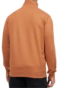 Homme portant un pull texturé orange-brun à manches longues et col montant, vu de dos, associé à un pantalon foncé.