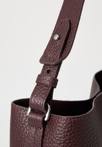 Sac en cuir bourgogne avec une surface texturée, bandoulière ajustable avec quincaillerie en argent, et une ouverture ronde en haut.
