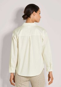 Helle gelbe Bluse mit einer Rückenfalte, langen Ärmeln und Knopfmanschetten, kombiniert mit gemusterten Hosen mit geometrischem Design.