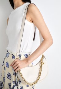 Borsa in pelle beige con forma curva e tracolla a catena dorata. La borsa è dotata di chiusura a zip e una texture liscia. Gonna con motivo floreale visibile.