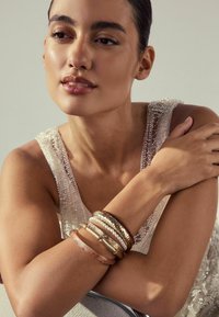 A model's arm features multiple bracelets: transparent acrylic, metallic, and pearl, with intricate designs and a neutral color palette.